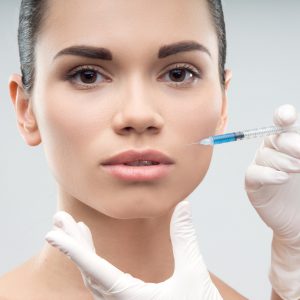 Beautiful young woman gets injection in her face by plastic surgeon, isolated on grey